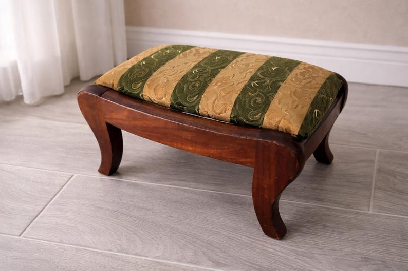 May include: A small, wooden footstool with a rectangular, cushioned top. The cushion features alternating vertical stripes of olive green and gold fabric. The curved legs are a dark brown color.