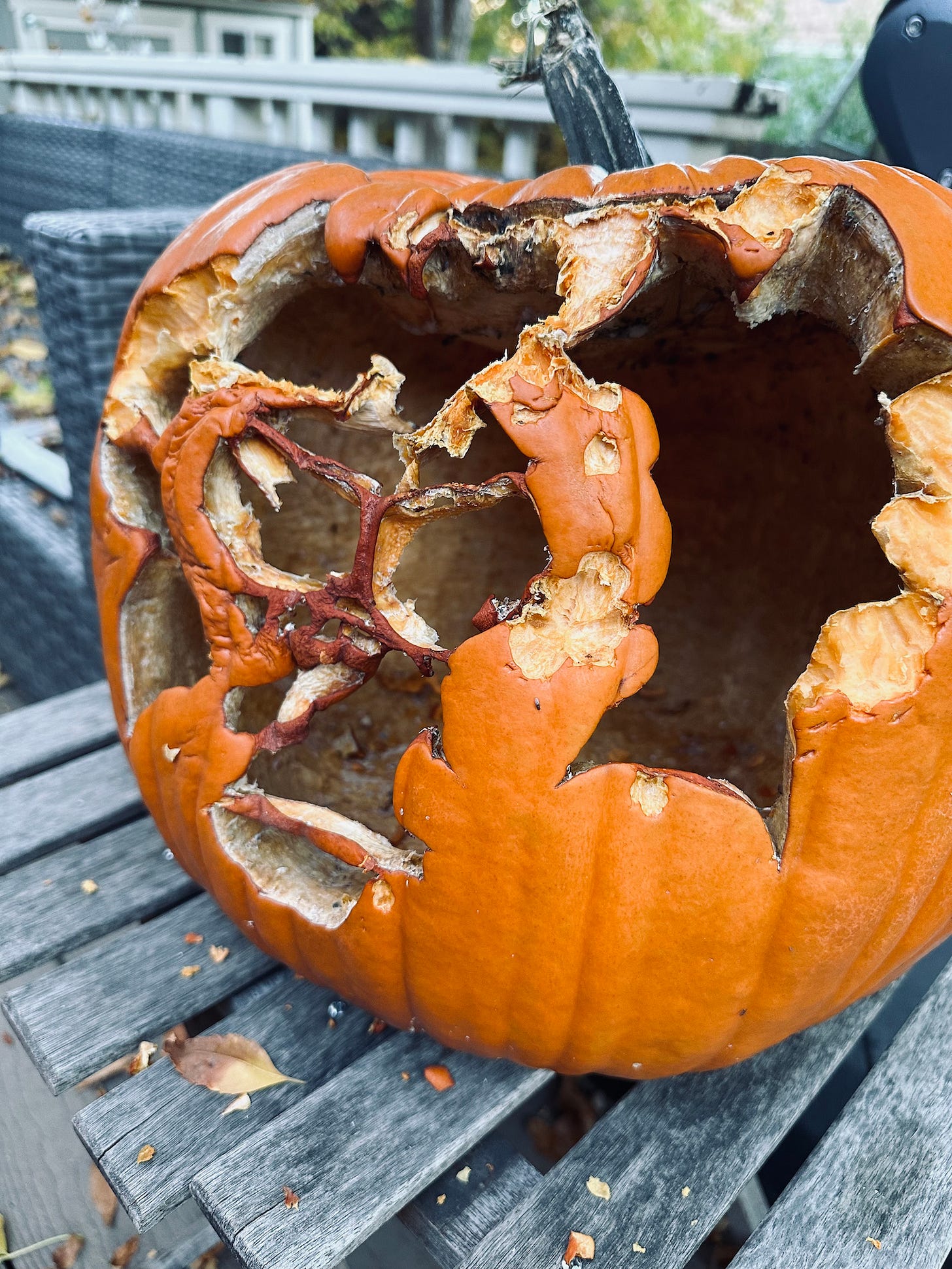 A largely rotted carved pumpkin sitting on an old, weathered wooden table by an outdoor couch. What was carved is no longer recognizable. 