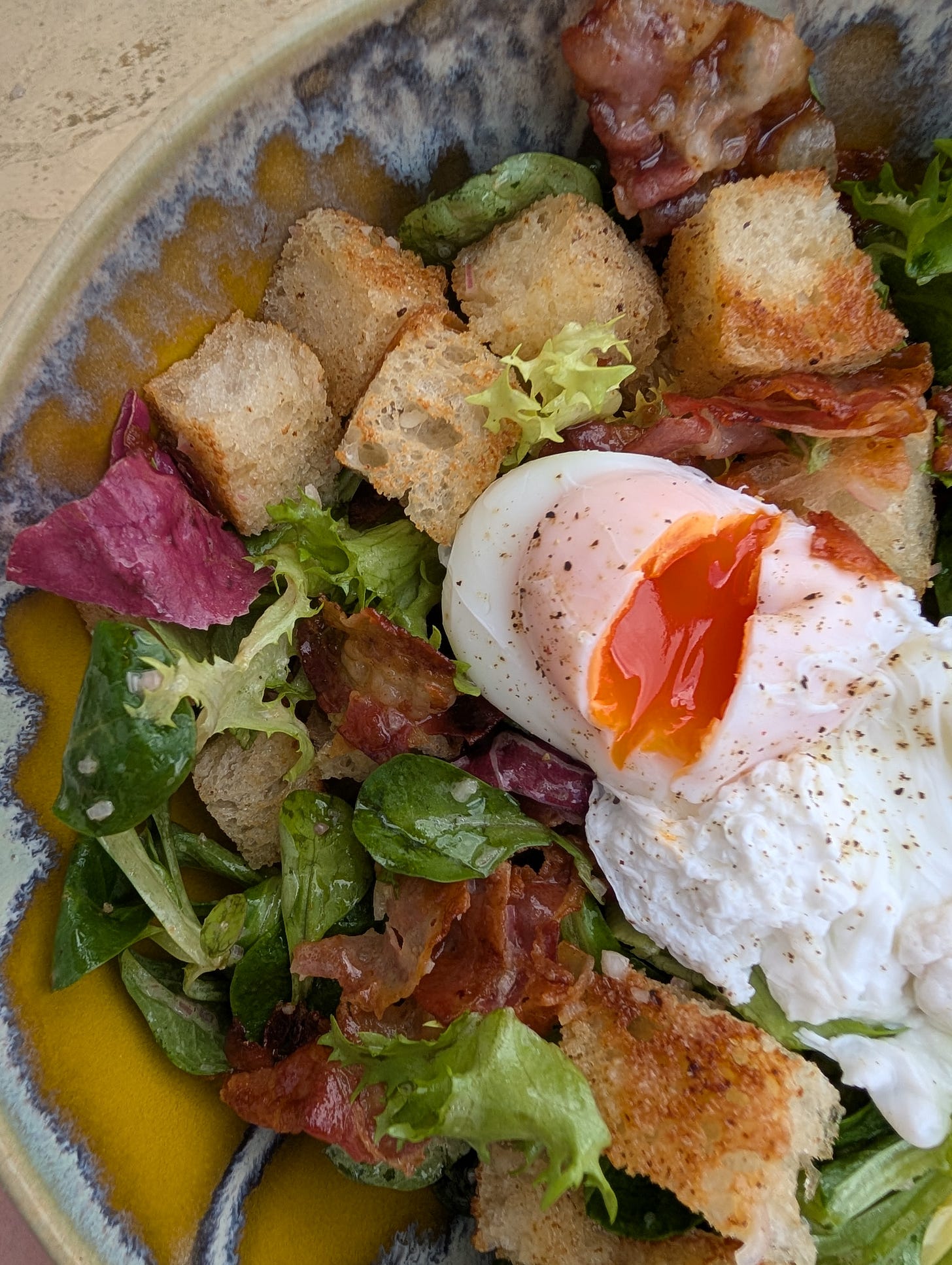 A close-up of a vibrant lyonnaise salad served in a rustic ceramic bowl. The dish features mixed leafy greens, crispy golden croutons, strips of cooked bacon, and a perfectly poached egg with a runny yolk just beginning to spill. The egg is seasoned with black pepper, adding a touch of spice to the fresh, savoury presentation.