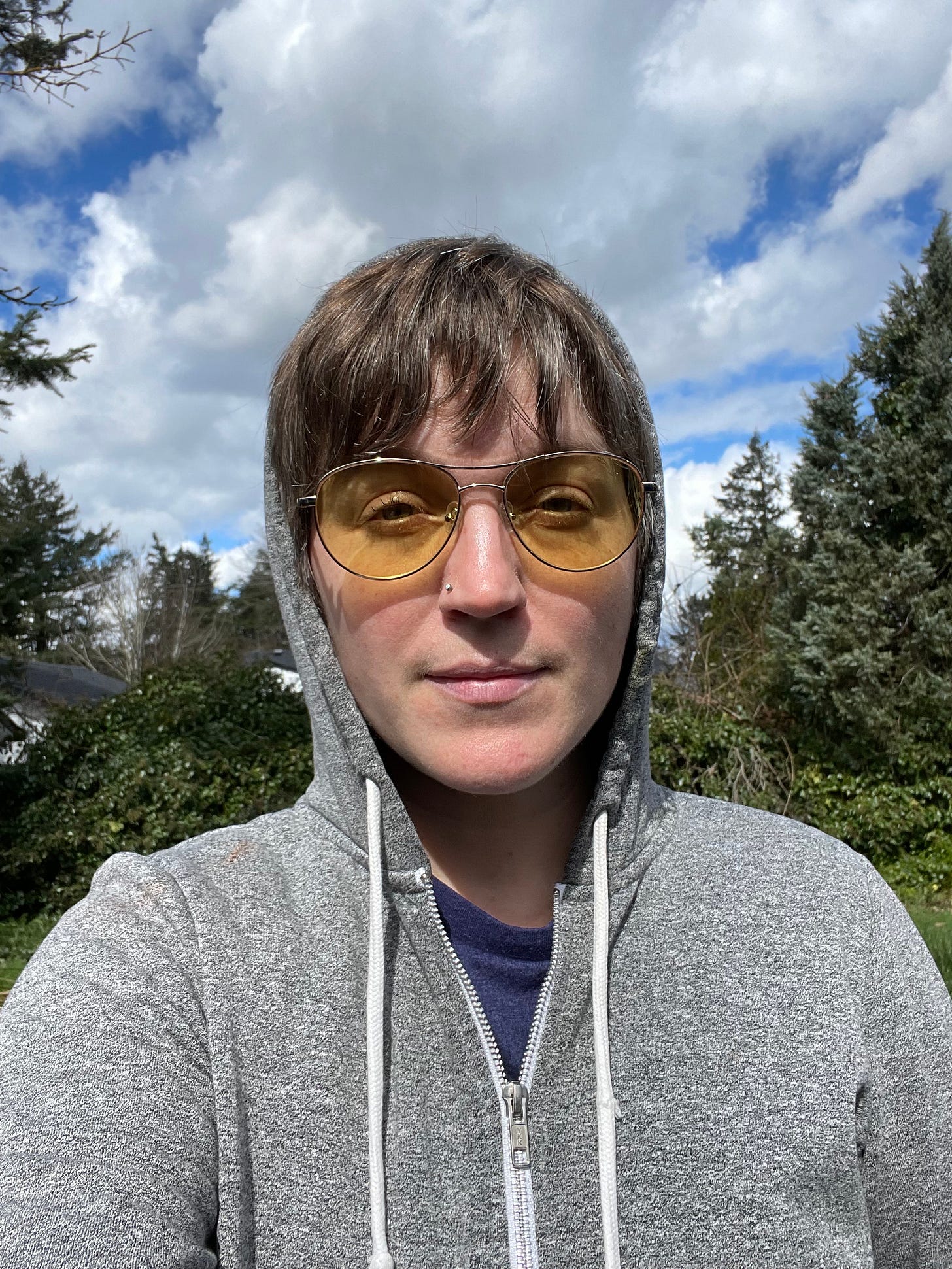 against a backdrop of blue sky and white clouds a middle aged person with short brown hair and a gray hoodie is wearing yelow tinted aviator glasses and is smiling slightly at the camera while squinting
