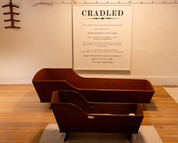 Two wooden cradles, one for an adult, the other for a child, are displayed on the floor of an exhibition space.