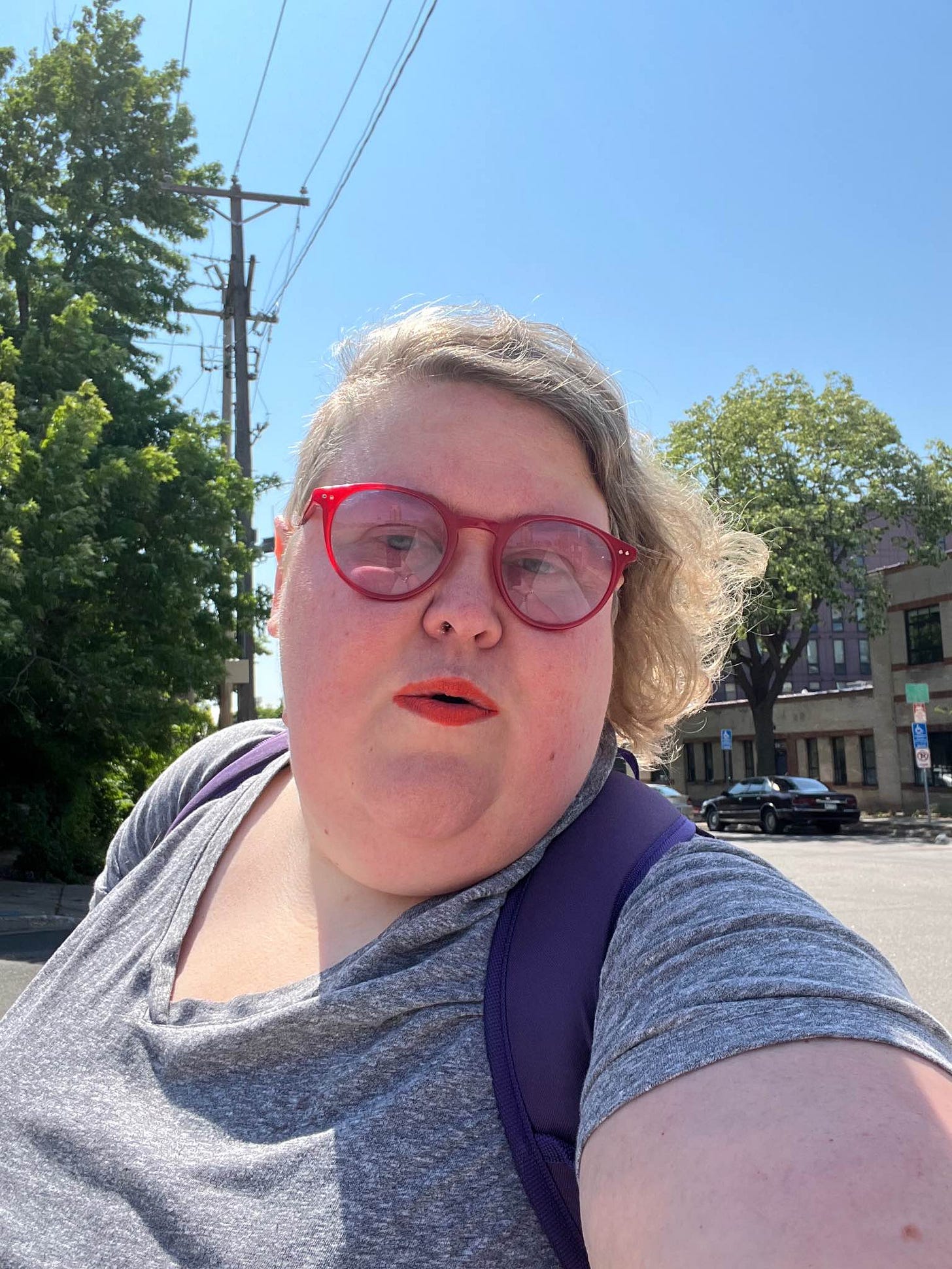 Me, Alison, a fat, white woman with red glasses, orange lipstick, and wearing a grey tshirt, under telephone lines with green trees, a street, a brick building, and a car in the background. My purple backpack straps are visible.
