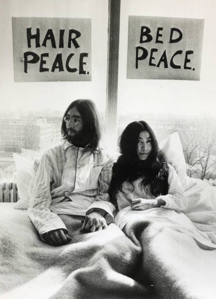iconic photo of John Lennon and Yoko Ono in bed on their honeymoon. John is on the left with a sign taped to a window behind his head that reads Hair Peace. Yoko, on the right, has a sign behind her head that reads Bed Peace