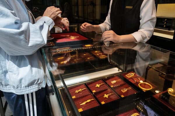 A person in a light blue jacket holds a gold bracelet over a red display tray. Another person in a black vest stands across a glass display case filled with gold jewelry.
