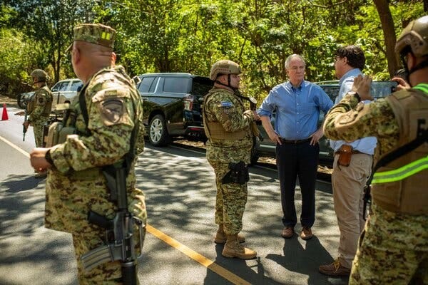 Senator Chris Van Hollen and Chris Newman, in collared shirts, speak with a military member in camouflage, as other officers stand nearby on a road.