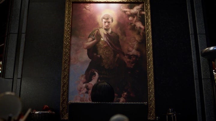 A grand, Renaissance-style oil painting of a younger Adrian Veidt (Ozymandias) in Roman armor, surrounded by cherubs. A dark silhouette of a person stares up at the portrait from below.