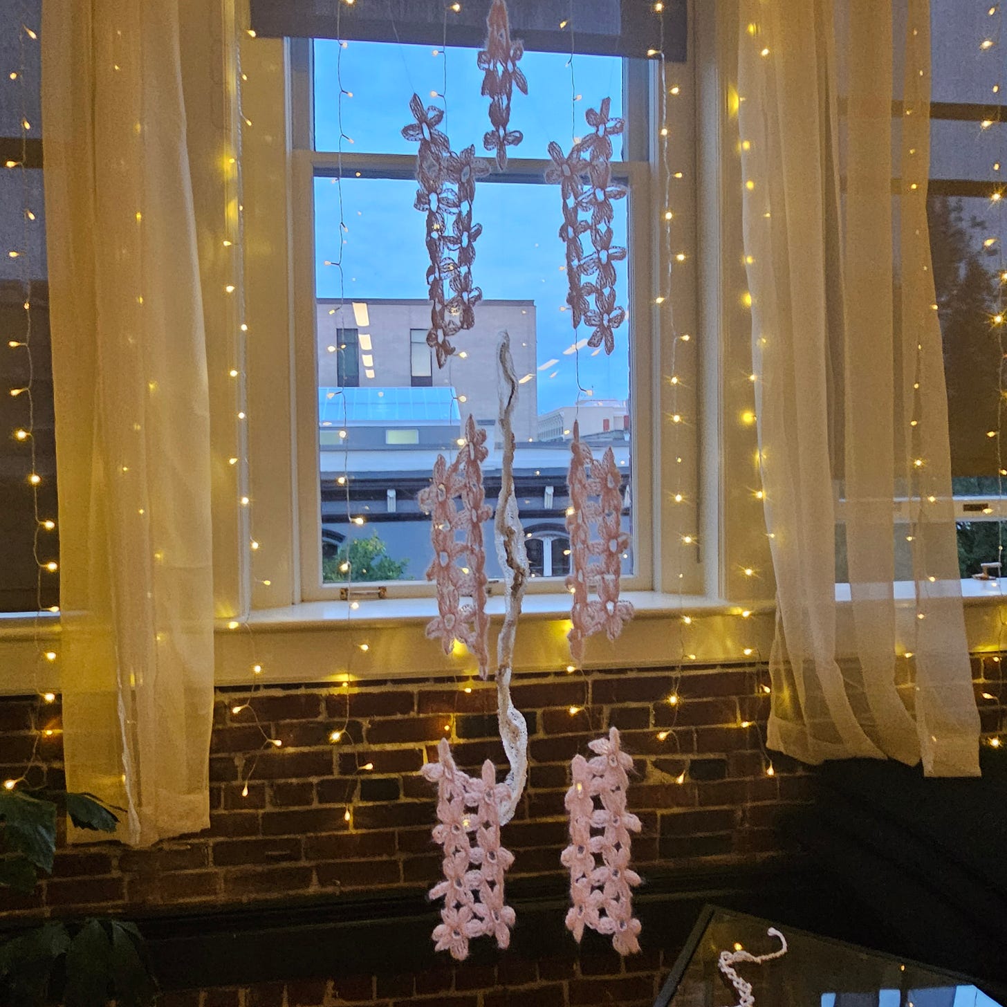  Pretty Pain hanging from the ceiling. It has invisible thread holding pink embroidered pieces together in front of a window illuminated with small fairy lights