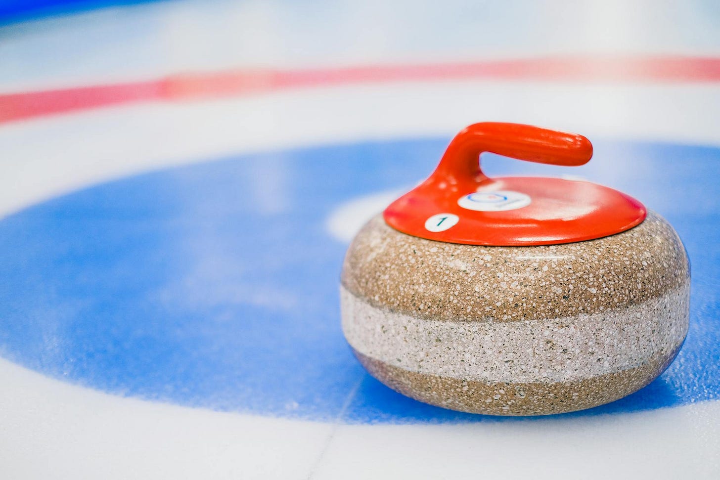 Primo piano di una pietra da curling (granito) con maniglia arancione, posizionata sulla superficie di ghiaccio di una pista da curling. La pietra si trova nella "casa", l'area bersaglio circolare, visibilmente sull'anello blu più esterno. La maniglia mostra il numero identificativo '1'.