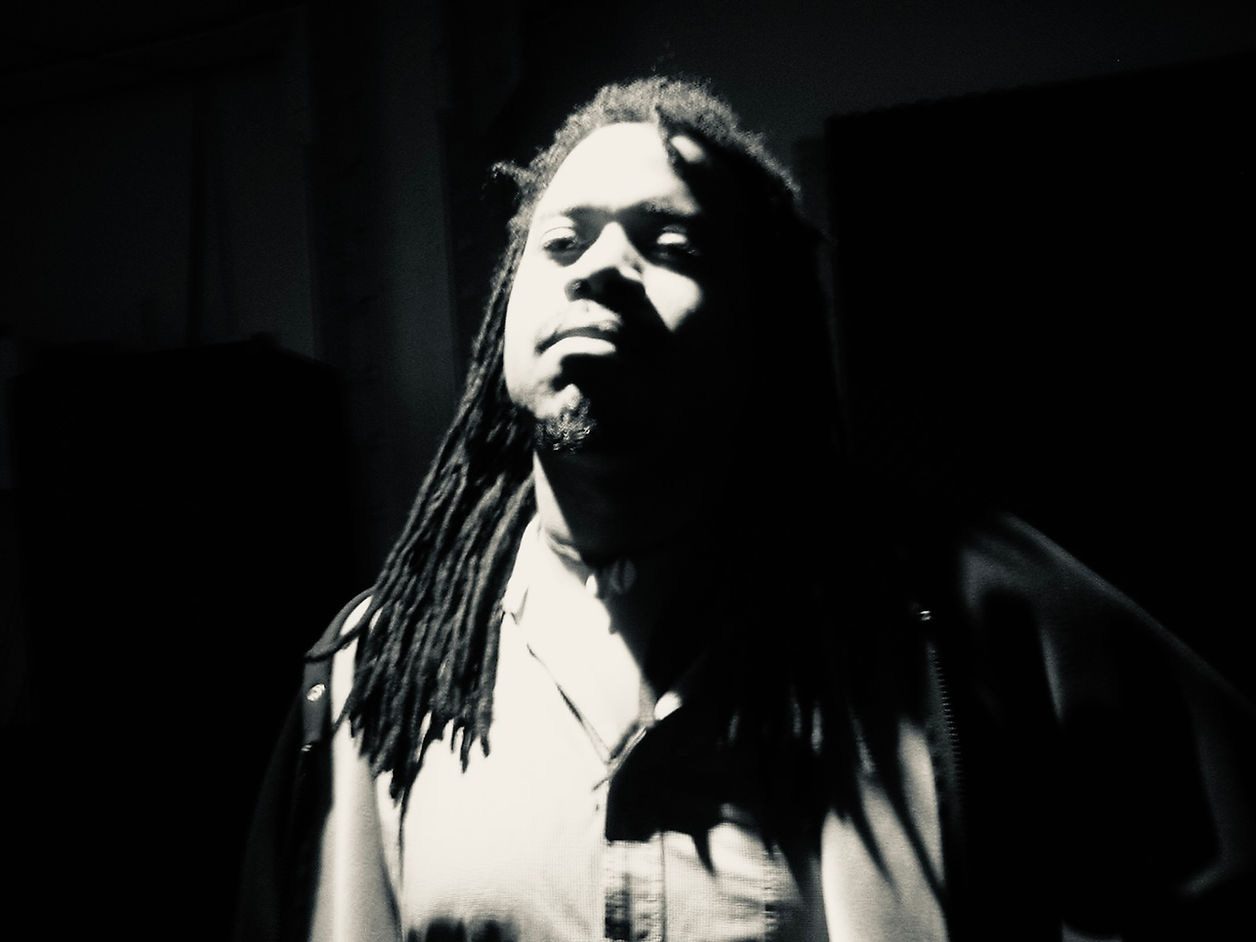 Person with dreadlocks in dim light, casting shadows. Dark background creates a dramatic, mysterious mood. Monochrome tones. 