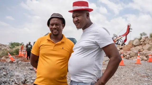 Mandla Charles and Mzwandile Mkwayi, the two community volunteers who descended into the abandoned mine, stand side by side near the top of the mine shaft. The one on the left is wearing a yellow T-shirt and the other is wearing a white T-shirt. Mandla Charles and Mzwandile Mkwayi, the two community volunteers who descended into the abandoned mine, stand side by side near the top of the mine shaft. The one on the left is wearing a yellow T-shirt and the other is wearing a white T-shirt.
