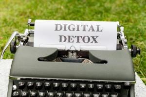 A typewriter on a table outside, with grass visible in the background. The typewriter has a piece of paper in it, with the words "digital detox" in black block capitals written on it. A typewriter on a table outside, with grass visible in the background. The typewriter has a piece of paper in it, with the words "digital detox" in black block capitals written on it.