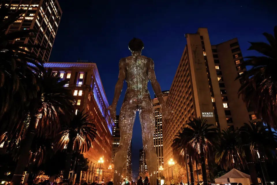 A towering illuminated sculpture of a nude female figure stands in a San Francisco plaza at night, viewed from behind, with palm trees and downtown buildings lining Market Street in the background.