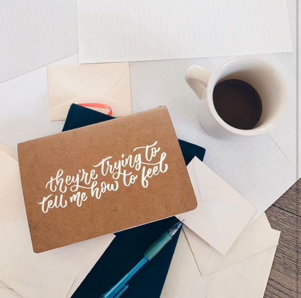A brown notebook on top of a pile of stationery with a cup of coffee beside it. The notebook has hand lettering in white script that says "they're trying to tell me how to feel".