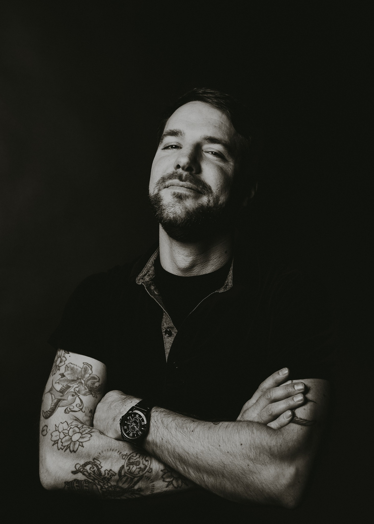 Black and white photo of man with tattoos. His arms are crossed and there's a smile on his face.