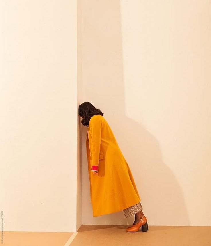This may contain: a woman in a yellow coat leaning against a white wall with her head on the corner