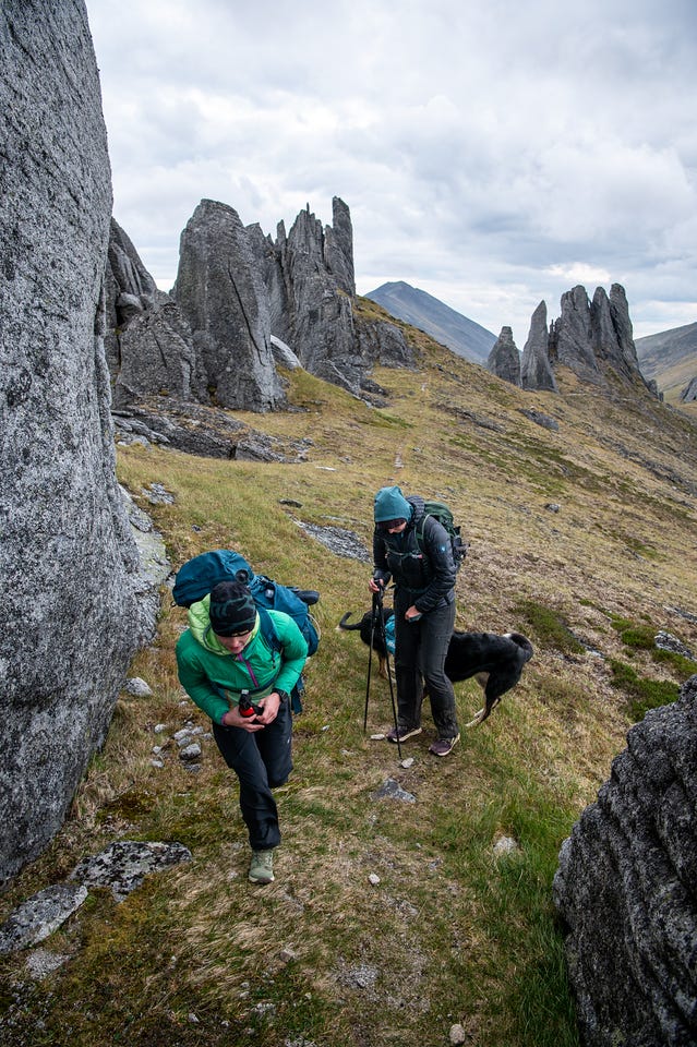 Two hikers with packs and a black-and-white dog follow a faint grassy path winding between towering granite pinnacles and fins, one hiker bent to the trail. A dark peak shows through a gap in the towers under a cloudy sky.