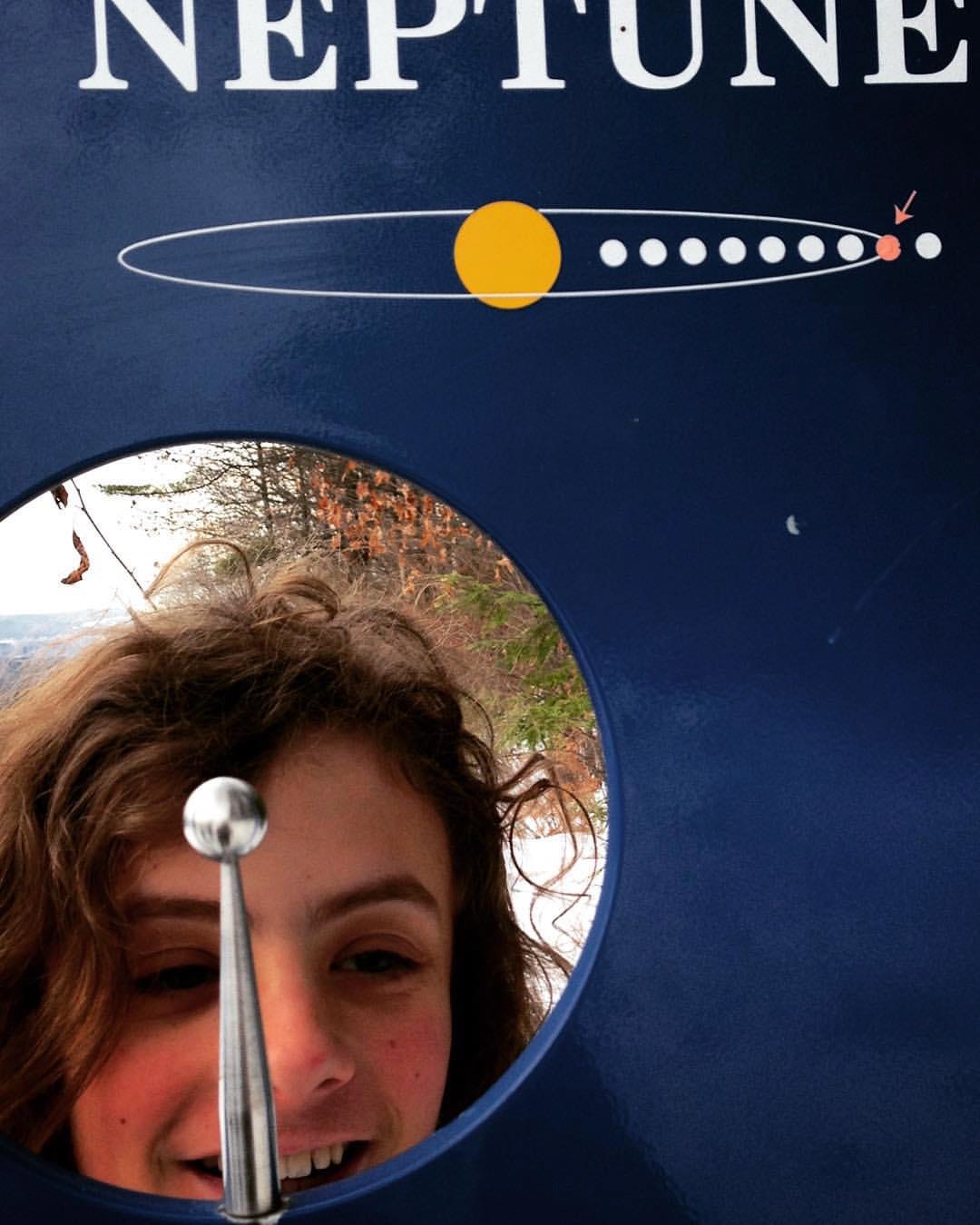an outdoor museum display says "neptune." through a hole in the display, the face of a teenager is vislbe. he has long curly hair and is smiling. an outdoor museum display says "neptune." through a hole in the display, the face of a teenager is vislbe. he has long curly hair and is smiling.