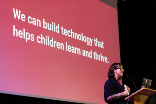 A person on stage stands before a slide with the sentence "We can build technology that helps children learn and thrive". A person on stage stands before a slide with the sentence "We can build technology that helps children learn and thrive".