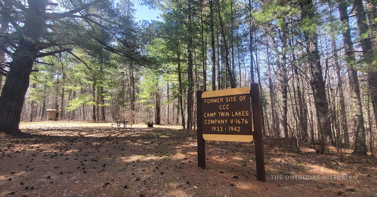 A horizontal wooden sign at Wanoka Campground in the Chequamegon National Forest. The sign marks the site of the former CCC Twin Lakes camp, surrounded by a lush forest of tall white and red pines on a bright, sunny spring day.