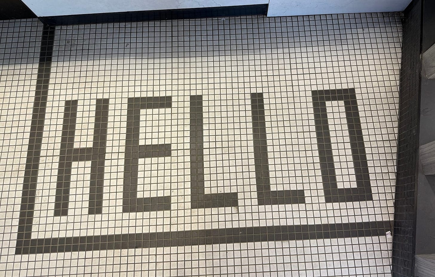 The image shows a tiled floor with the word “HELLO” spelled out in large, black, capital letters. The tiles are small and square, with a white background and black lines forming the letters and a border around the word. The word “HELLO” is centered within the border.
