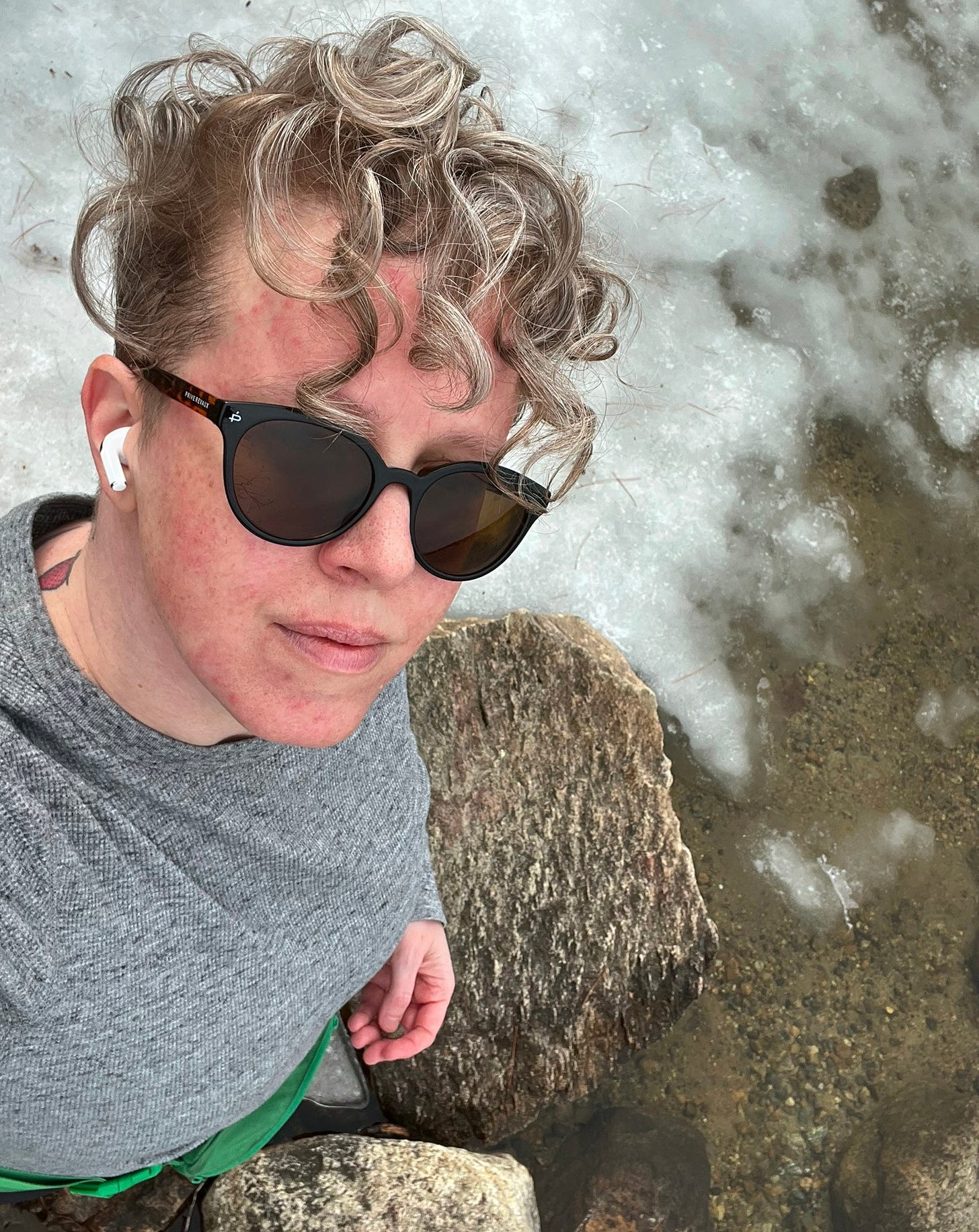 a white trans nonbinary person with short curls and sunglasses looks up from the edge of a partially icy lakeside