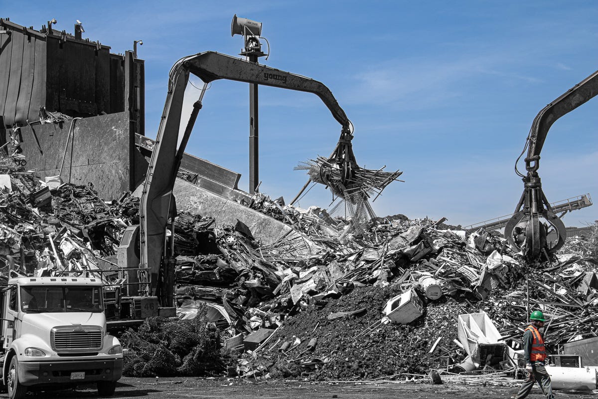 Wide shot of three excavators with claws lifting metal in a junkyard, one worker in safety gear walking at bottom right. Wide shot of three excavators with claws lifting metal in a junkyard, one worker in safety gear walking at bottom right.