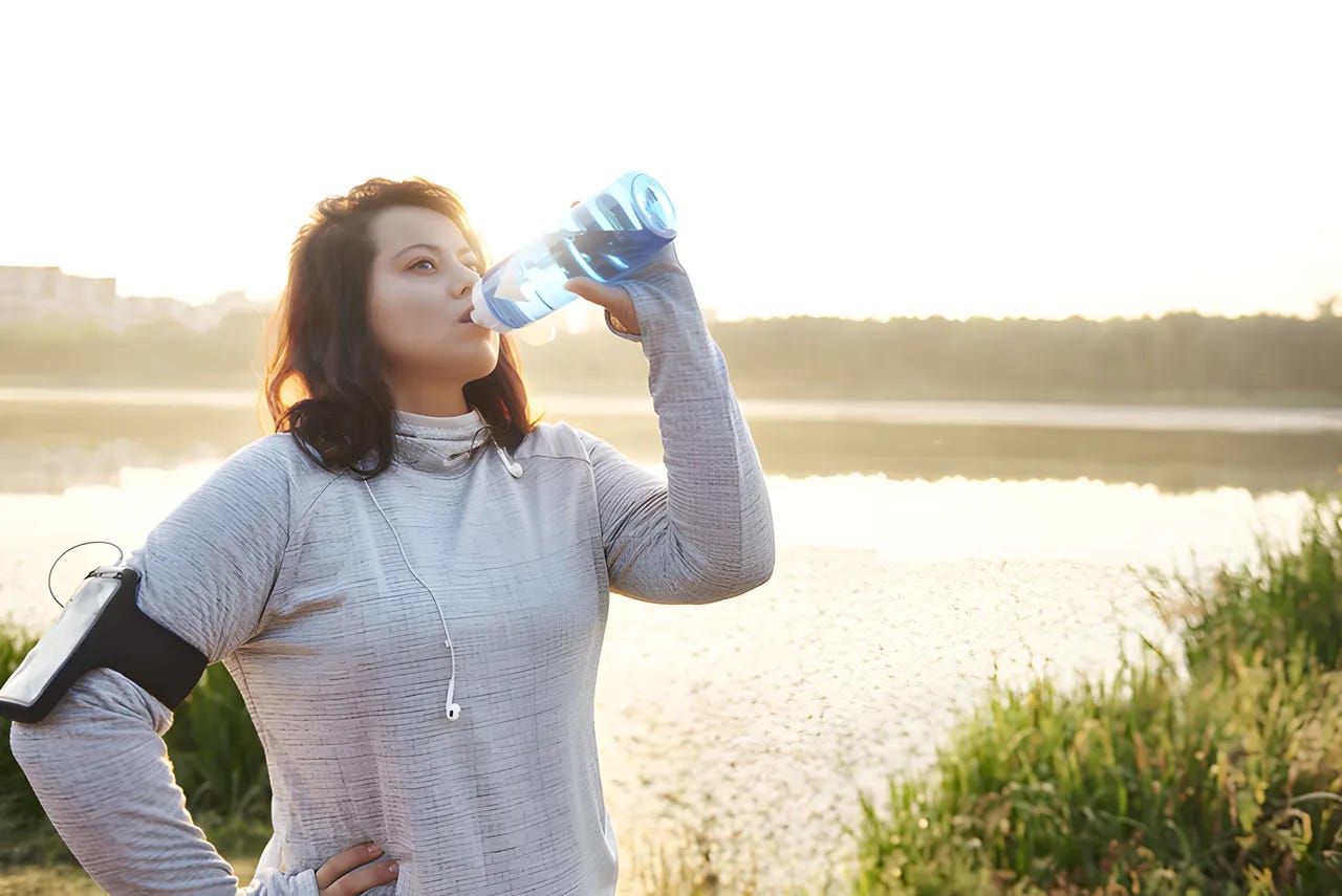 Woman drinking out of a water bottle after finishing a run by a pond.