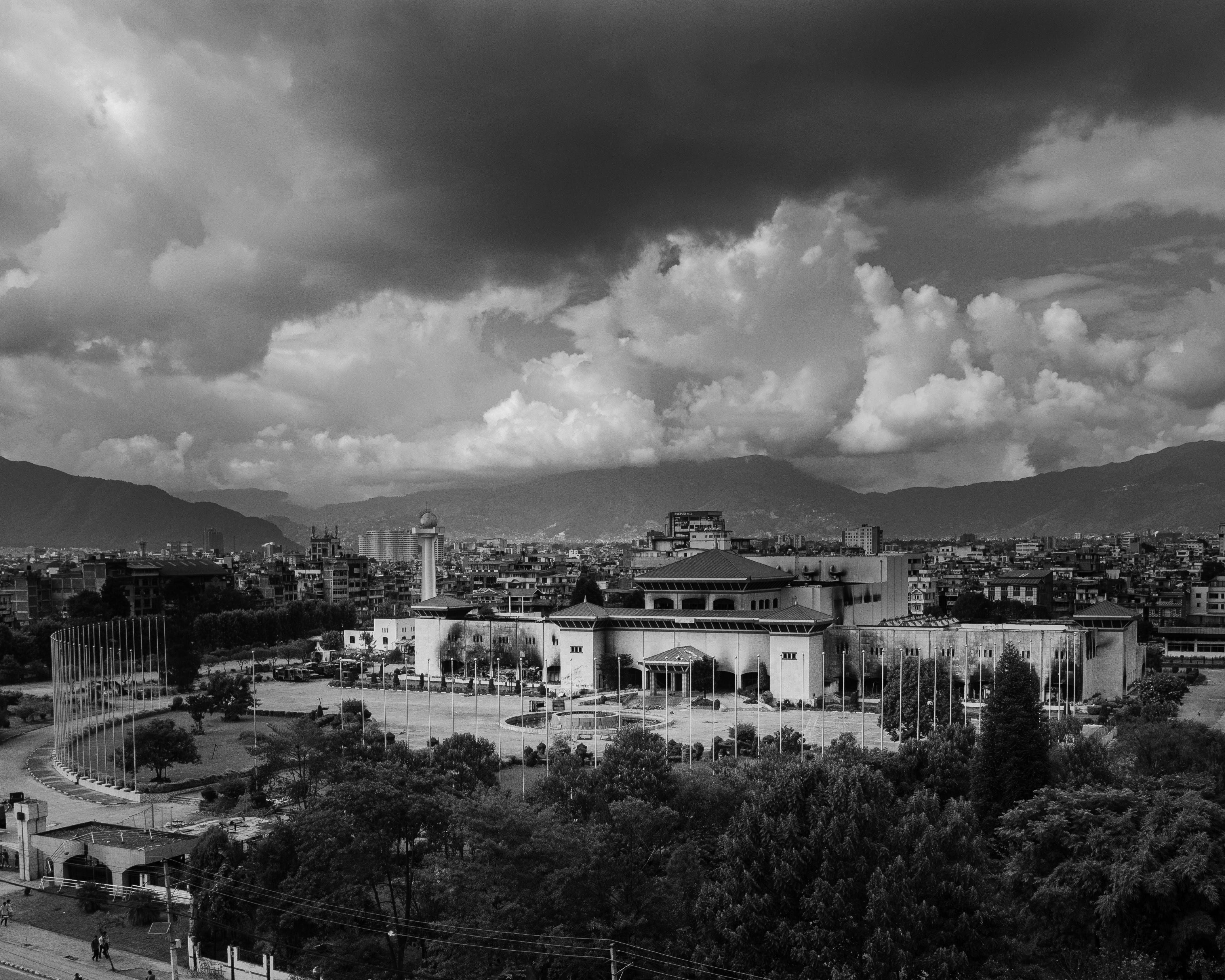 A black-and-white photograph of the Sansad Bhawan (Parliament Building) in Kathmandu, Nepal, taken after the GenZ protest. The dramatic sky shows a striking contrast, heavy dark clouds on one side and bright, illuminated clouds on the other; symbolizing uncertainty, struggle, and hope for a new dawn.Photo: Bijay Chaurasia Photography, Wikimedia Commons, License CC-BY-SA 4.0
