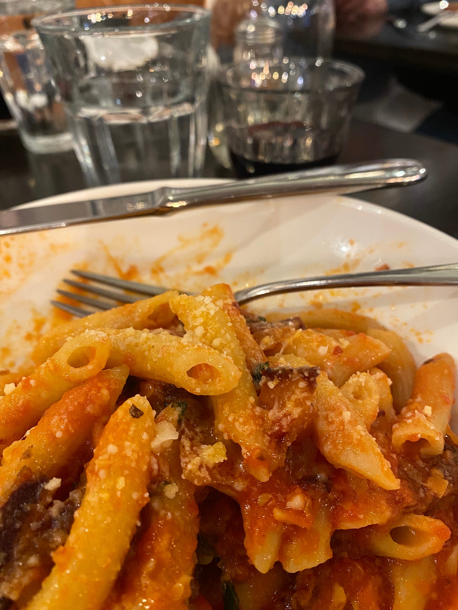 A close-up photo of a bowl of penne in a red sauce sprinkled with parmesan. In the background are a short glass of wine and a taller glass of water.