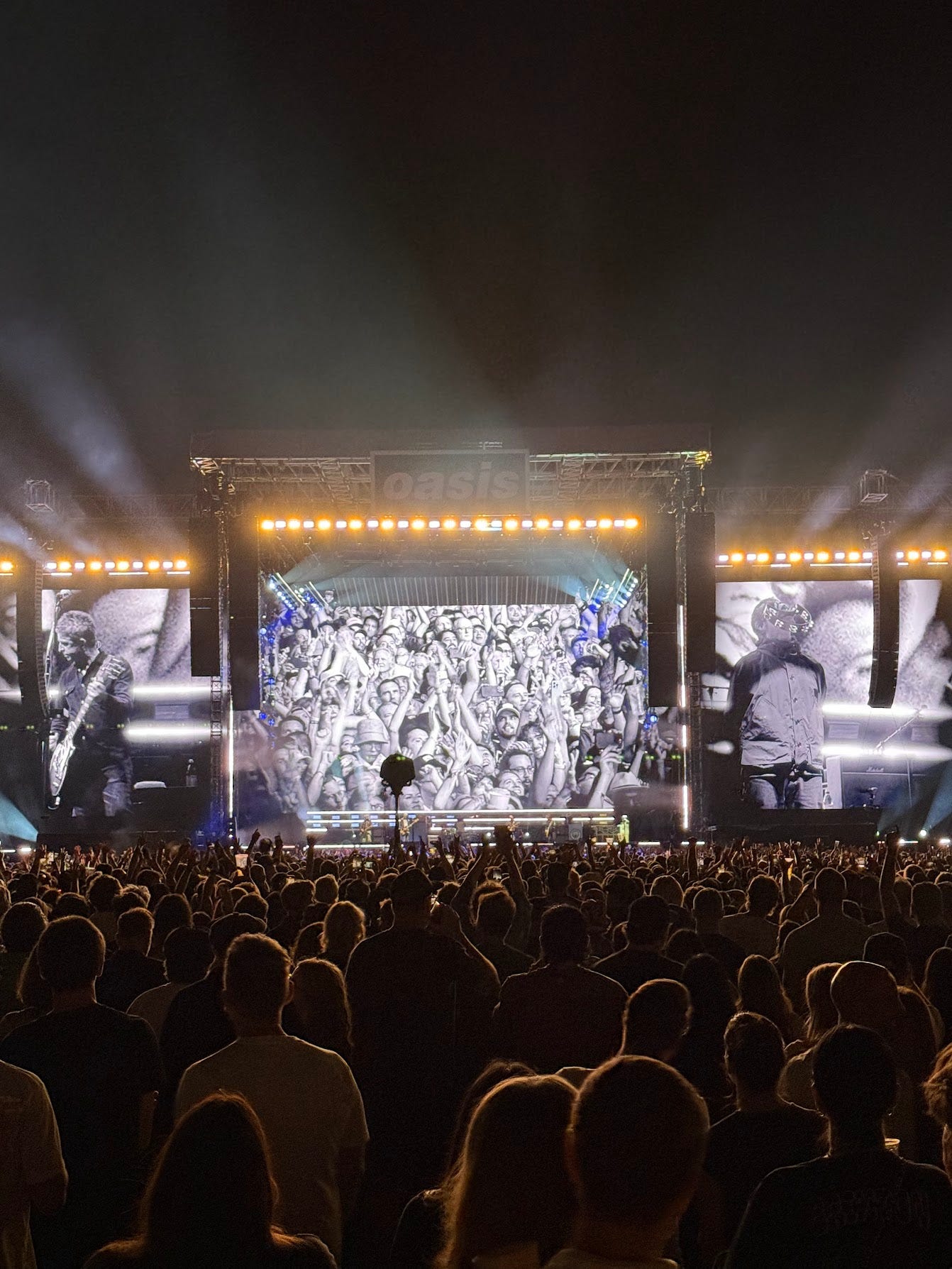 A photograph taken from the back of the field-level crowd of Oasis’ performance on Sept. 6 2025 in Pasadena. Three huge video screens are visible on the stage, with Noel Gallagher playing guitar on the left-hand one, Liam Gallagher holding a tambourine in his mouth in the right-hand one, and on the center one a crowd shot of attendees at the concert waving their arms. The speaker stacks are visible hanging from scaffolds in front the screens. A row of orange and blue stage lights shines toward the crowd from above all the screens, casting radiant beams through the smoke. At the top of the center scaffold, over the band, is an “Oasis” logo sign. The band themselves are dwarfed by the state setup, and barely visible as tiny figures under the center screen. All the audience members are rim-lit in orange by the stage lights.