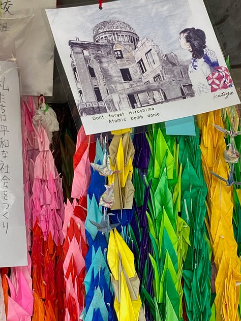 Photo of the Children's Peace Memorial, a tall concrete structure with a crane on top. Colorful strings of folded cranes. Museum display with tiny cranes foleded by Sasaki.