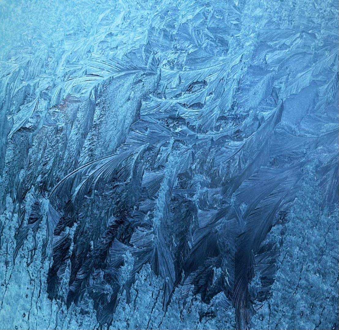 A close-up of frost patterns on a car window in winter. Delicate, feather-like ice formations spread across the glass in shades of blue, resembling frozen waves or intricate brushstrokes, revealing nature’s quiet artistry