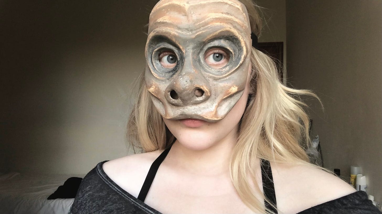 Liv in college wearing a Commedia dell'Arte mask she made. Chest up, she is looking at the camera, expressionless.