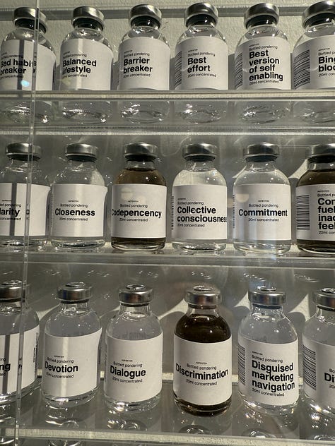 Art installation titled 'Bottled Pondering' by Aepenton, on display at the Design Museum Denmark. The piece features a wall-mounted grid of small glass vials, each labeled with a different abstract or emotional concept, such as 'Inner Critic Softener' or 'Courage Enhancer.' The vials are arranged in neat rows within a transparent display case, evoking the look of a scientific archive or apothecary cabinet of thoughts and feelings.