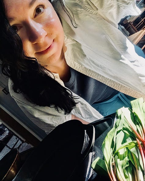 A person with black hair in a white shirt leans over a kitchen counter full of of ramps, a tomato plant in a cage perches next to a pink air fryer inside a kitchen, an orange cat rests his chin on a couch and looks annoyed