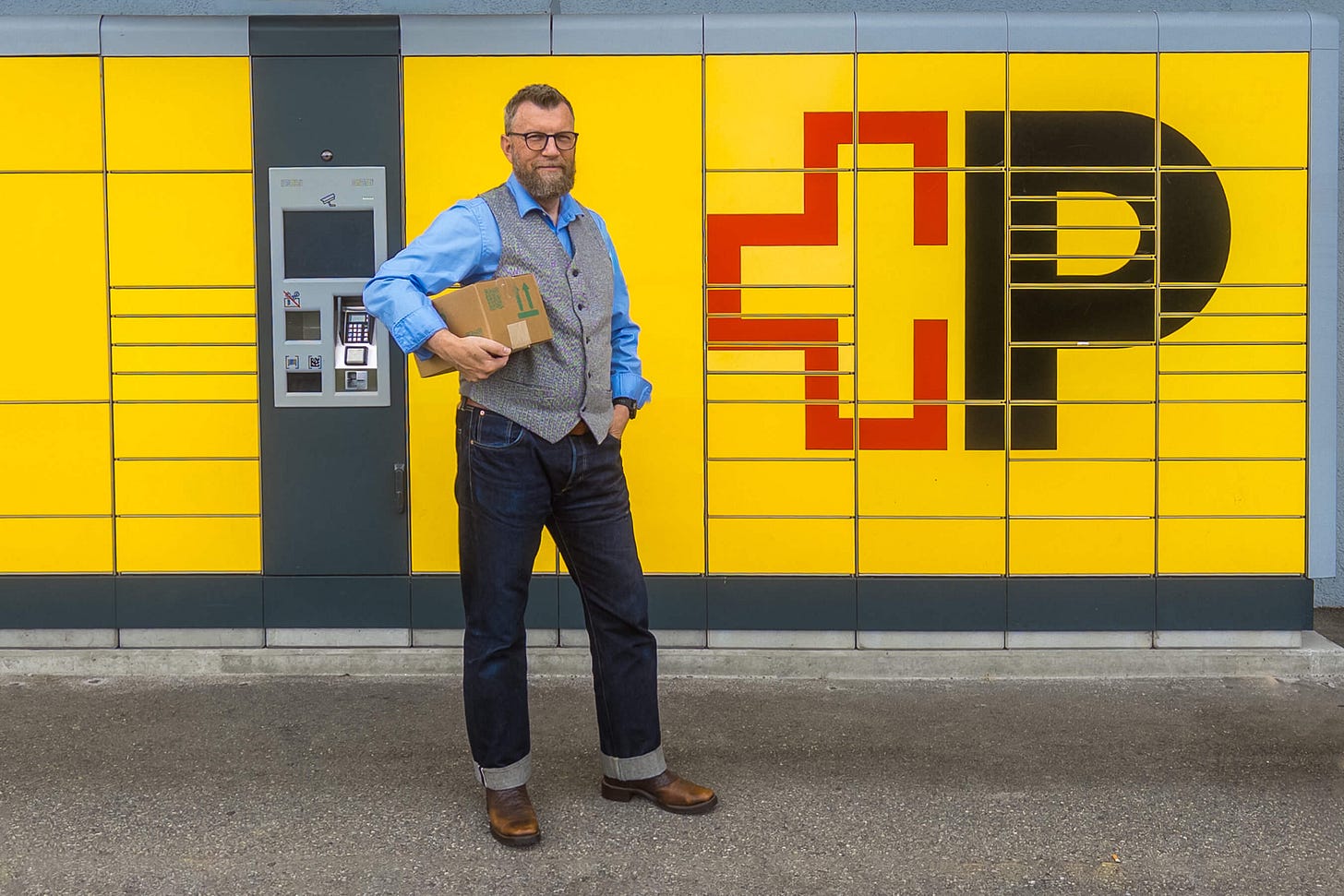 Jürg Vollmer steht vor einer gelben Paketstation der Schweizerischen Post. Er trägt eine Brille, blaues Hemd, graue Weste, Jeans sowie braune Cowboy-Stiefel und blickt in die Kamera. Unter dem Arm trägt er ein Paket. Hinter ihm ist das grosse schwarze P-Logo der Schweizer Post mit rotem Posthorn zu sehen. Jürg Vollmer steht vor einer gelben Paketstation der Schweizerischen Post. Er trägt eine Brille, blaues Hemd, graue Weste, Jeans sowie braune Cowboy-Stiefel und blickt in die Kamera. Unter dem Arm trägt er ein Paket. Hinter ihm ist das grosse schwarze P-Logo der Schweizer Post mit rotem Posthorn zu sehen.