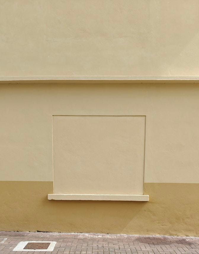 Una facciata di un edificio quasi minimalista e simmetrica, divisa in tre sezioni orizzontali e due colori complementari. La parte superiore e il centro sono di un colore giallo crema o beige chiaro uniforme. C'è una cornice o una sottile sporgenza orizzontale che separa la sezione superiore dal centro. Al centro, c'è un grande riquadro rettangolare incassato che simula una finestra o una nicchia murata, anch'esso dello stesso colore giallo crema. Sotto questo riquadro si trova un davanzale stretto e sporgente. La sezione inferiore del muro è di un giallo ocra o marrone chiaro più scuro, che crea un contrasto. La parte inferiore dell'immagine mostra un marciapiede di mattoni rossi e una striscia di vernice bianca che forma quadrati. La luce è uniforme, probabilmente diurna.
