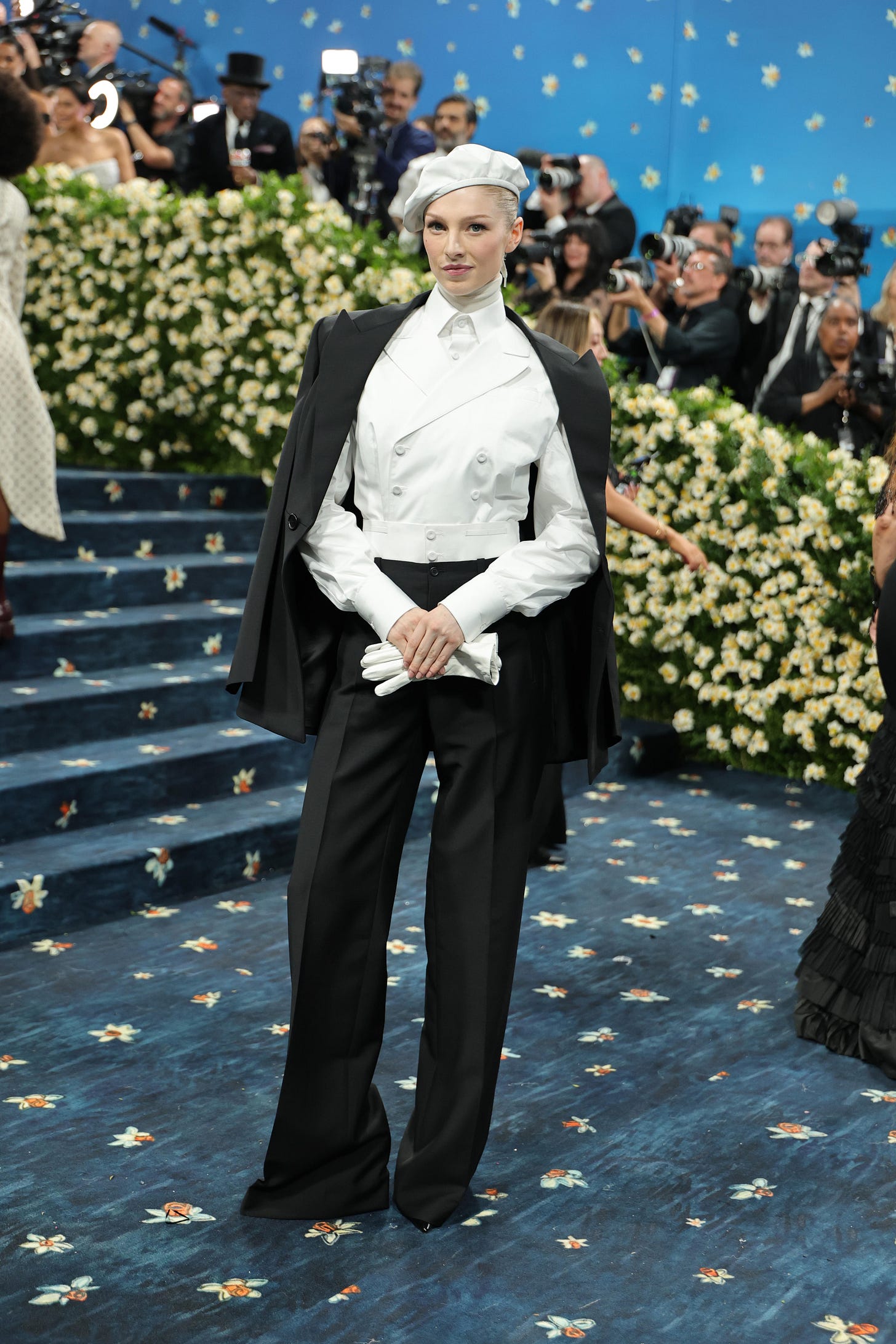 Hunter Schafer on the blue carpet in a white tailored suit top, with a black cloak, black pants, and a white beret