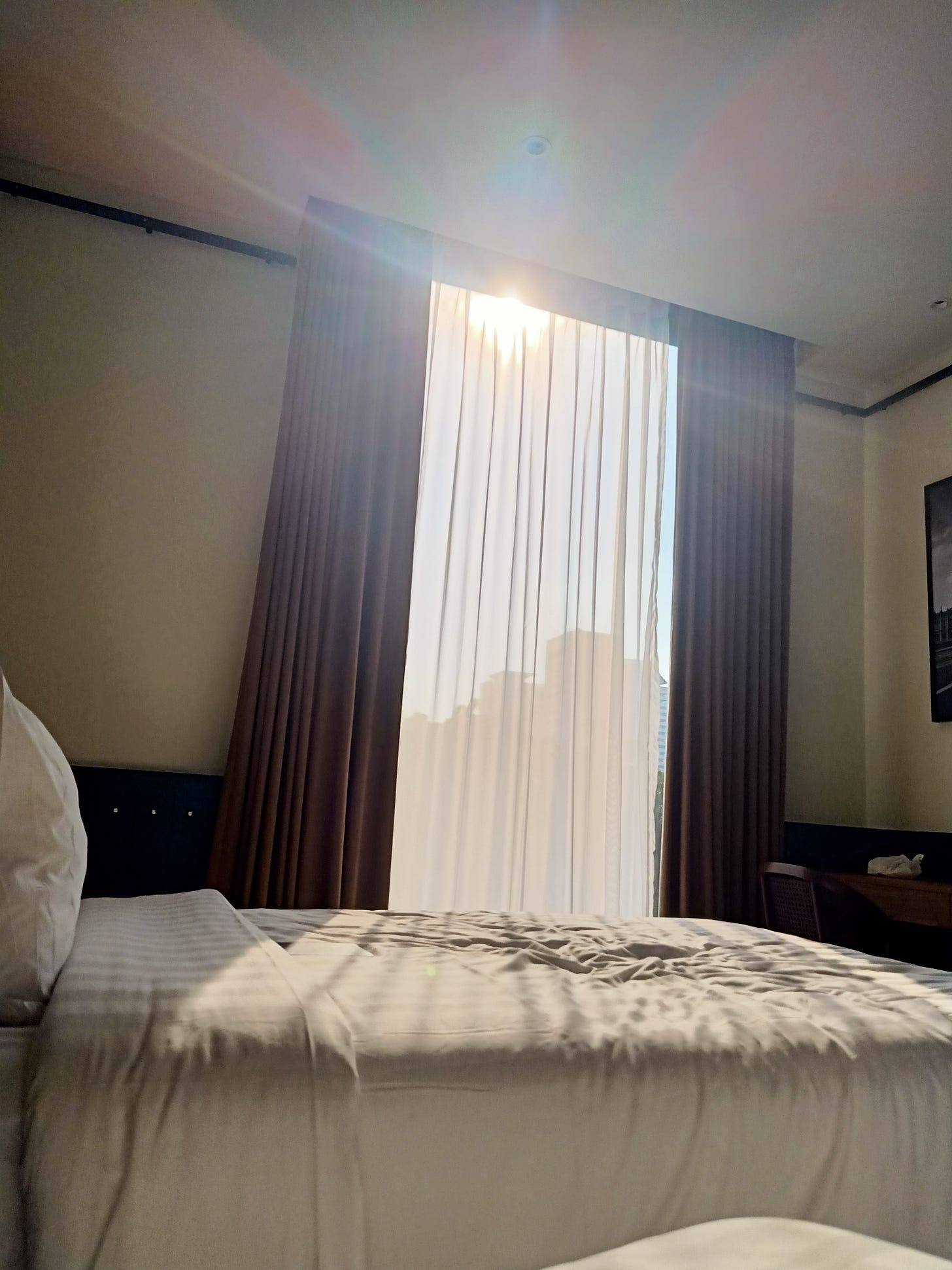 a white sheet covered hotel bed with crinkled sheets where someone has sat and a window behind with sunlight shining through the sheer white curtain