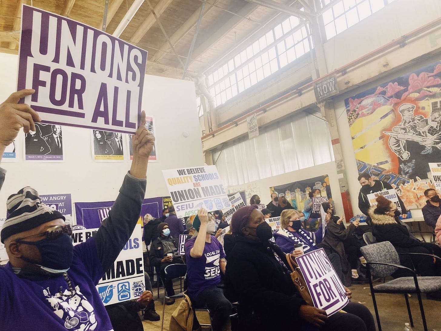 a crowd of people sitting wearing purple hold signs saying "unions for all" and "we believe quality schools are union made"