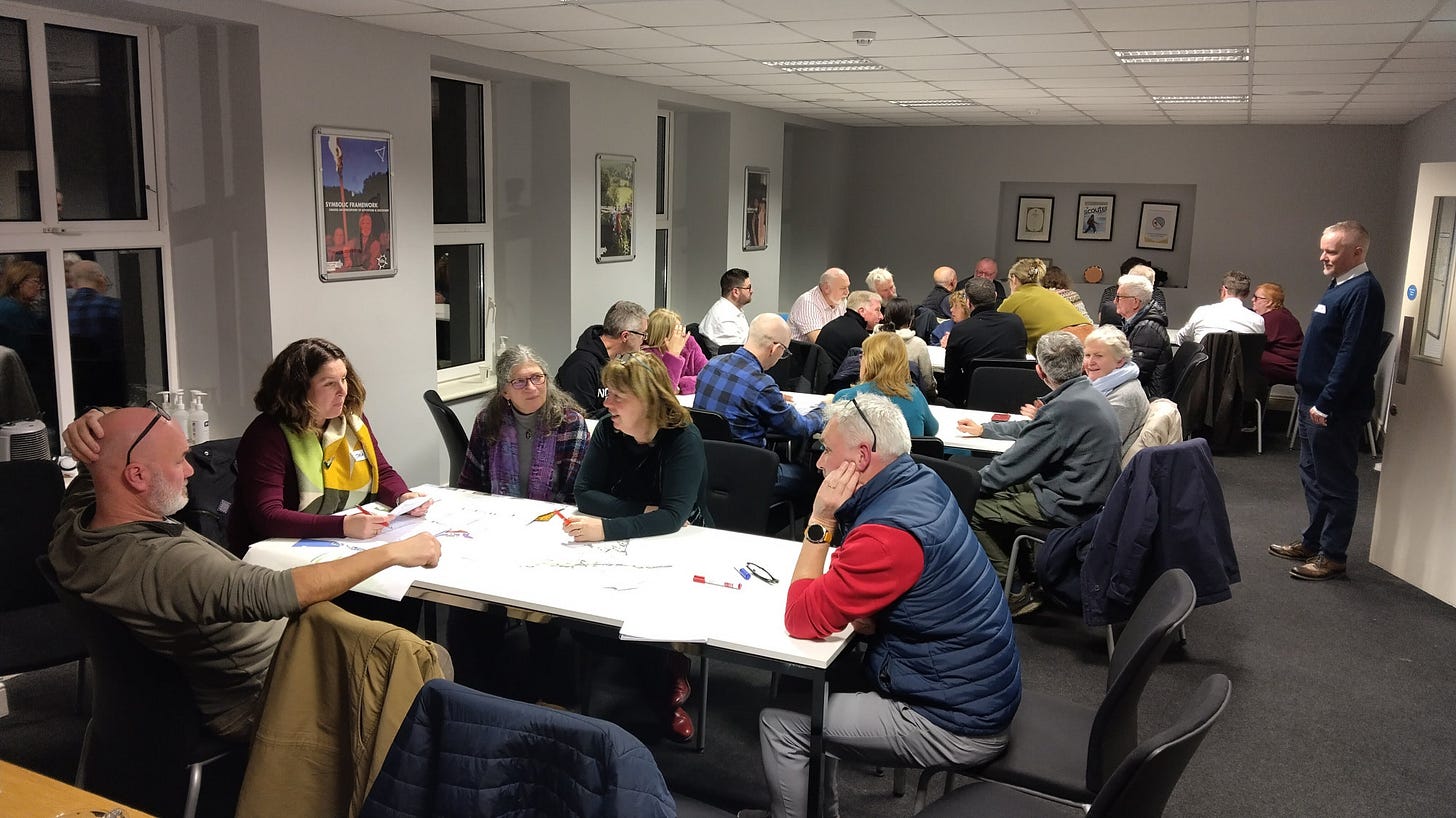 Members of the public enagaged in small-group conversations at tables in Kilcully scout hall.