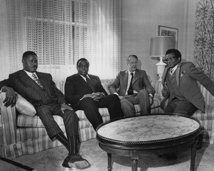 Four men in suits sitting on couches around a coffee table in a lounge setting. Four men in suits sitting on couches around a coffee table in a lounge setting.