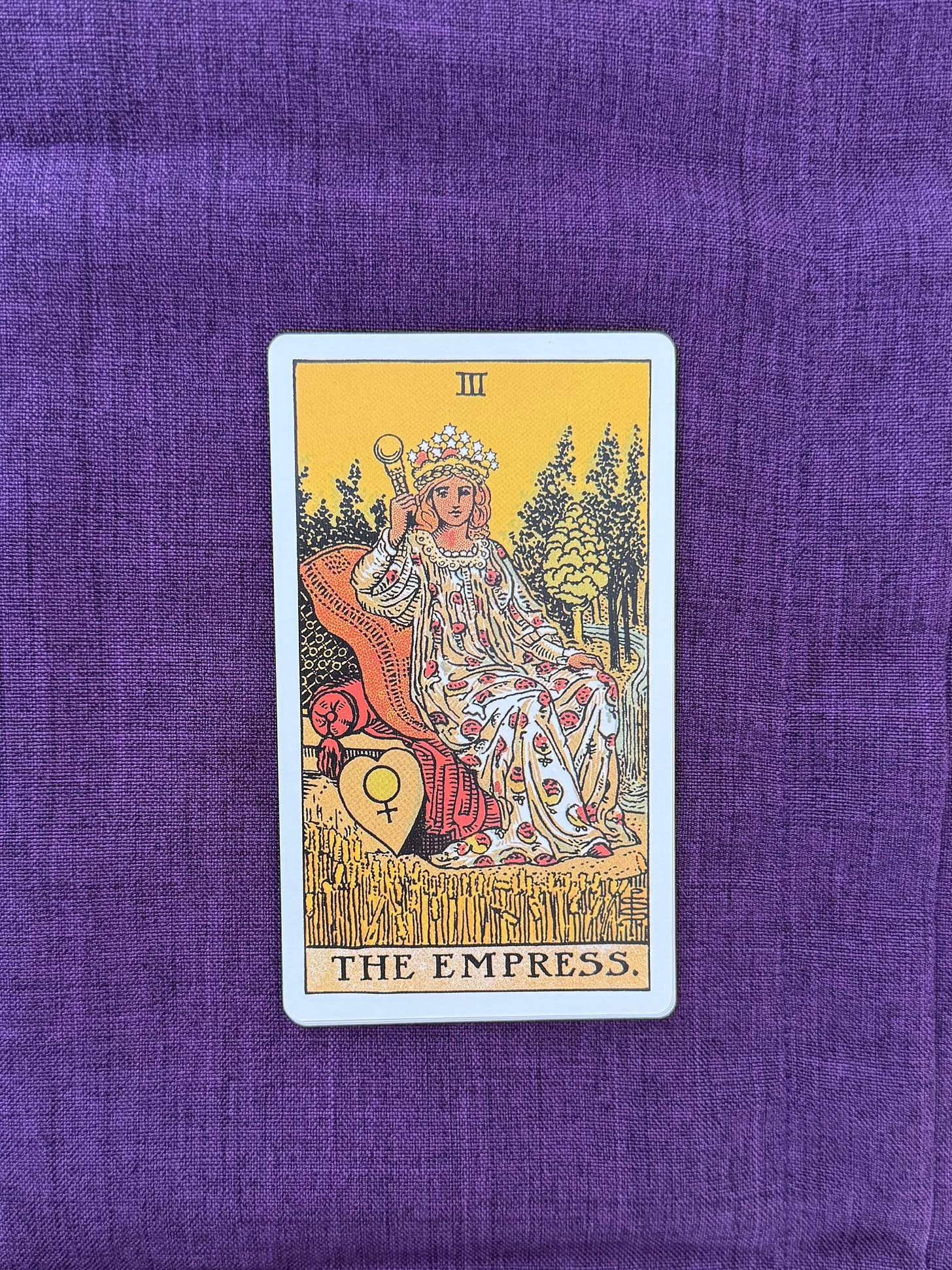 A seated figure wearing a crown and flowing robe sits on a cushioned throne surrounded by trees and wheat. The figure holds a scepter in one hand and rests the other hand on the arm of the throne. A shield decorated with the symbol of Venus rests beside the throne. A stream flows through the background toward distant trees and hills.