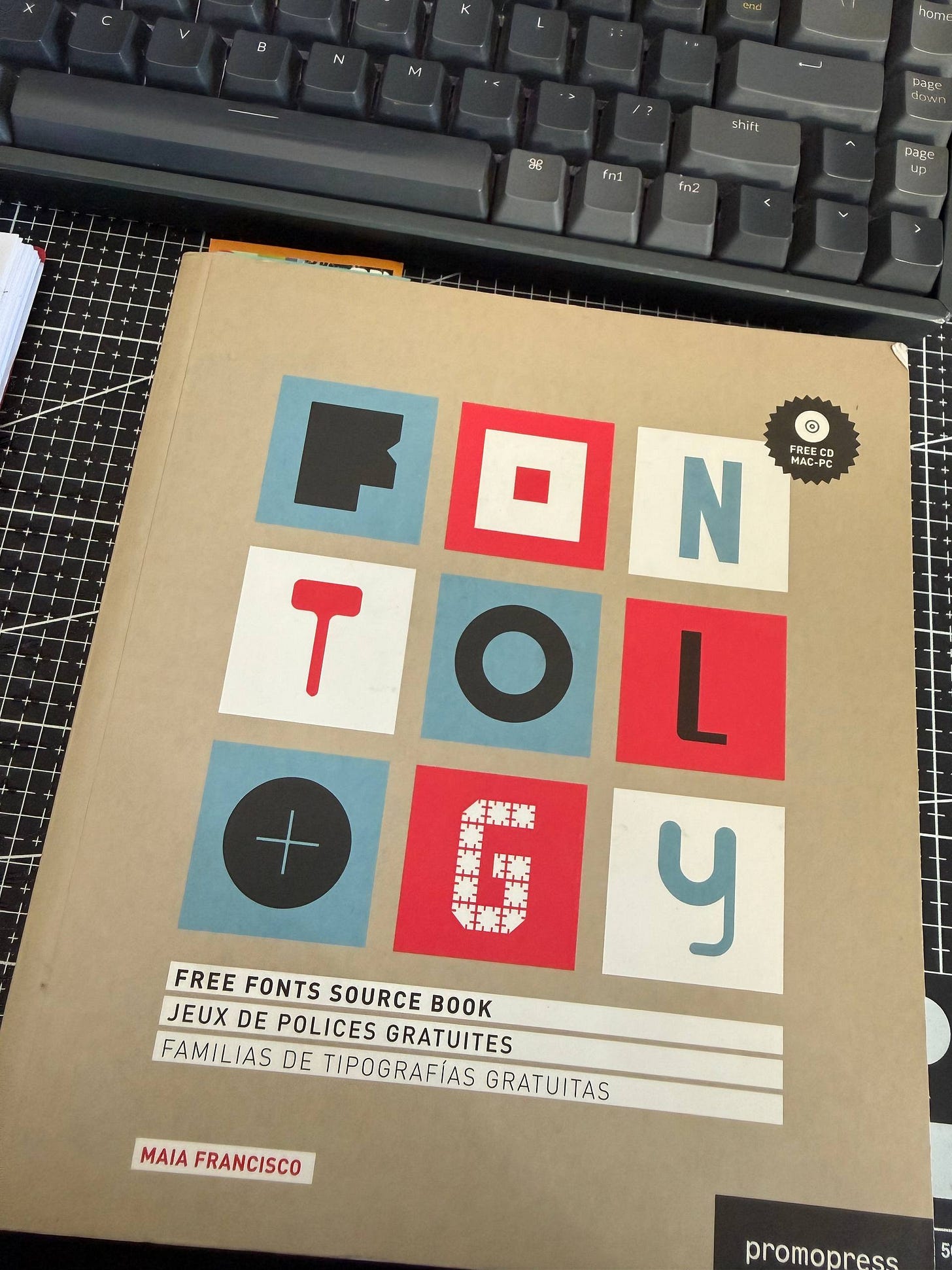 Portada del libro "Fontology: Free Fonts Source Book" de Maia Francisco, con un diseño de cuadrícula de letras coloridas, ubicado sobre un escritorio frente a un teclado mecánico negro.