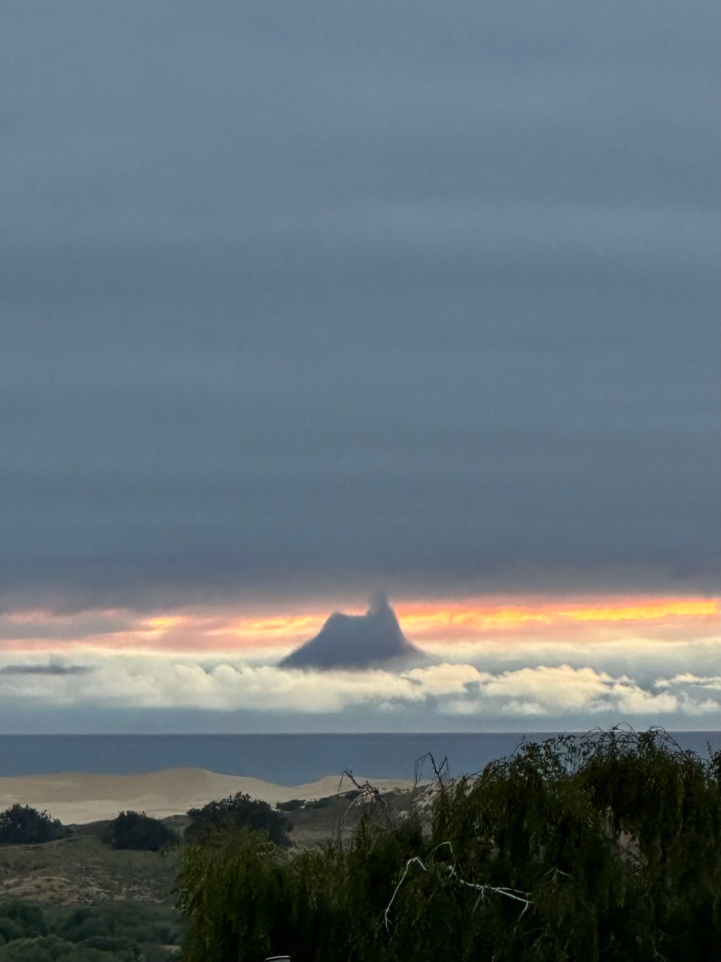 A close-up of a coastal landscape under a thick, brooding sky. A bright, narrow band of orange and yellow light pierces the horizon over a dark ocean. In the center of the light, a dark, jagged cloud formation rises upward, resembling a steep mountain peak. Pale sand dunes and deep green shrubs fill the foreground.