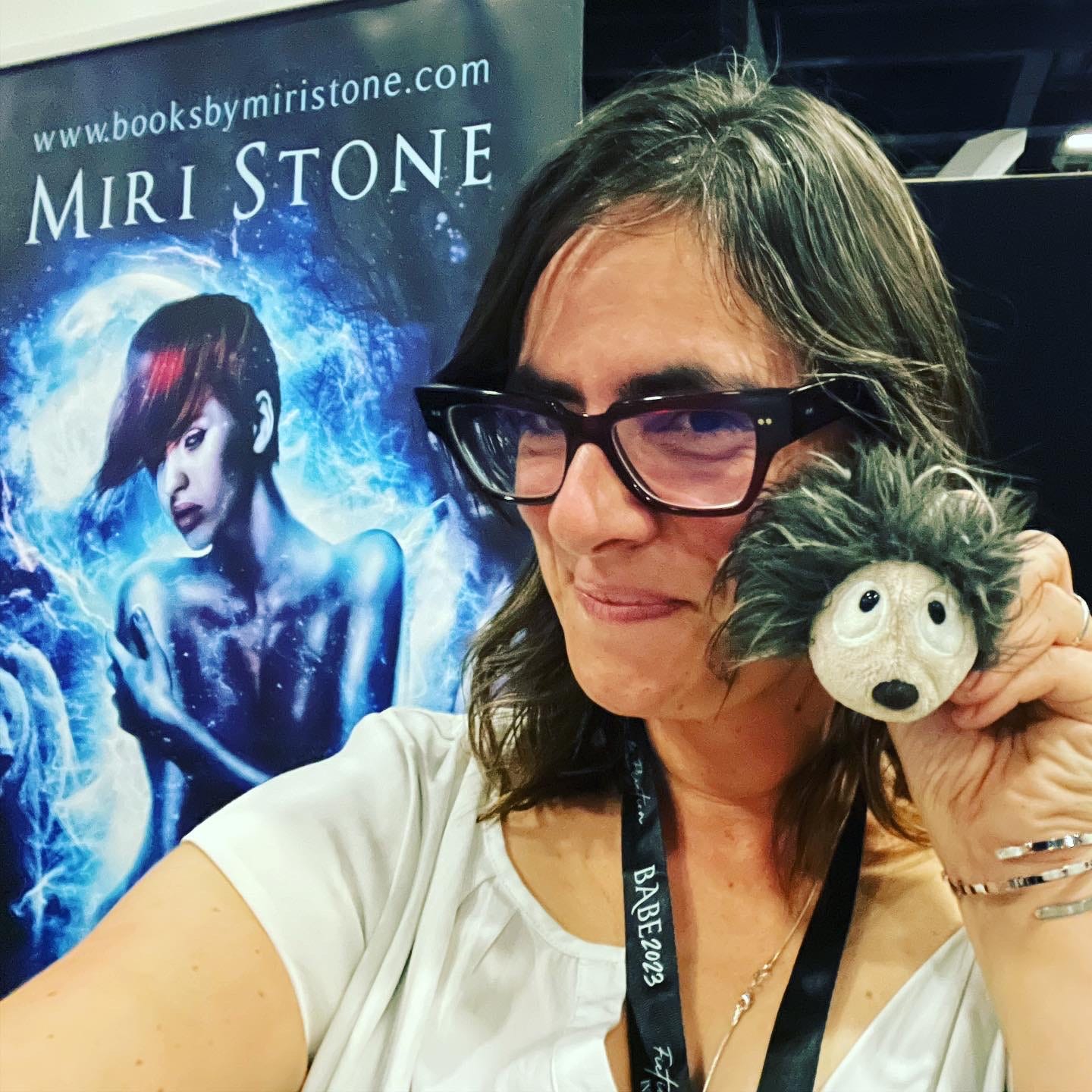 Miri Stone at a book signing with her Phoenix Crew author banner in the background and cuddled up to her hedgehog mascot, PINS, named after a favorite character in the Phoenix Crew series.