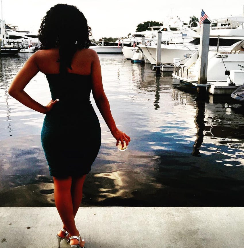 Woman standing on a marina dock looking out at boats and water while holding a drink.