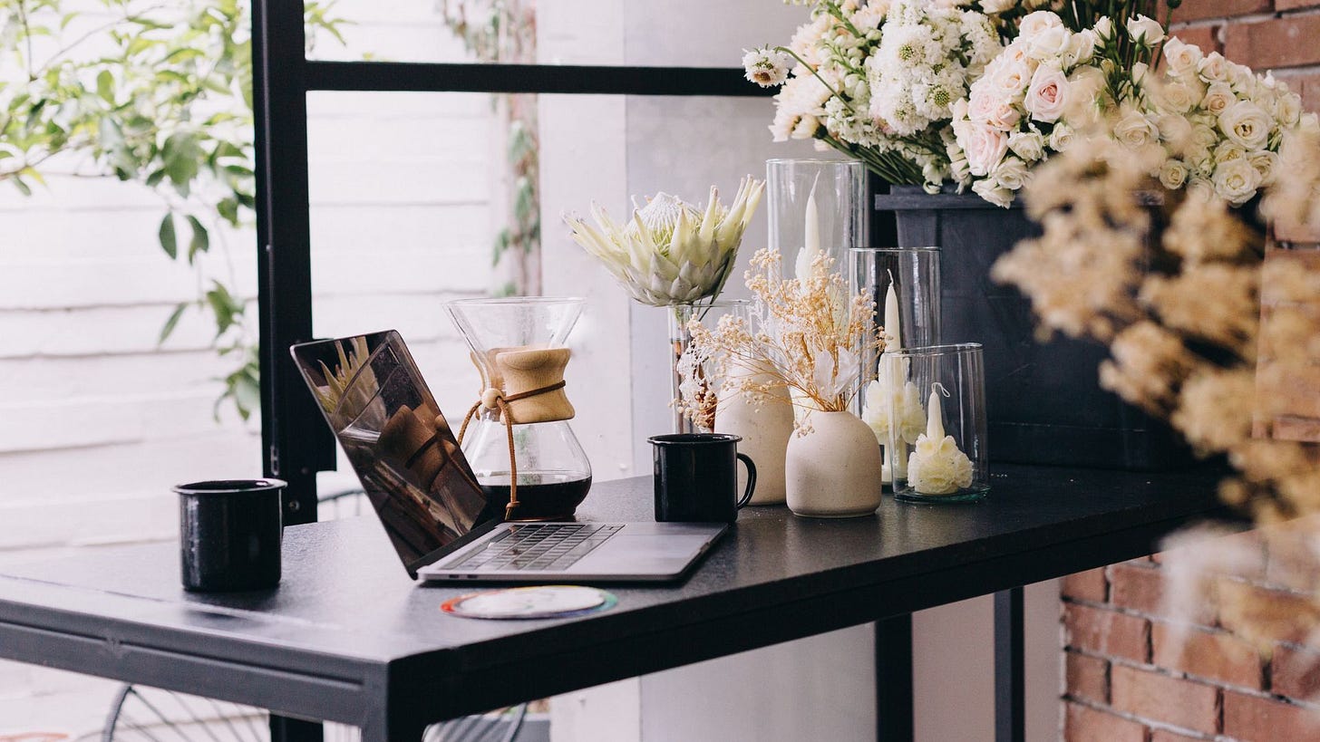 black, sleek desk facing a window next to an exposed brick wall with a laptop, coffe, carafe, and candles on the desk.