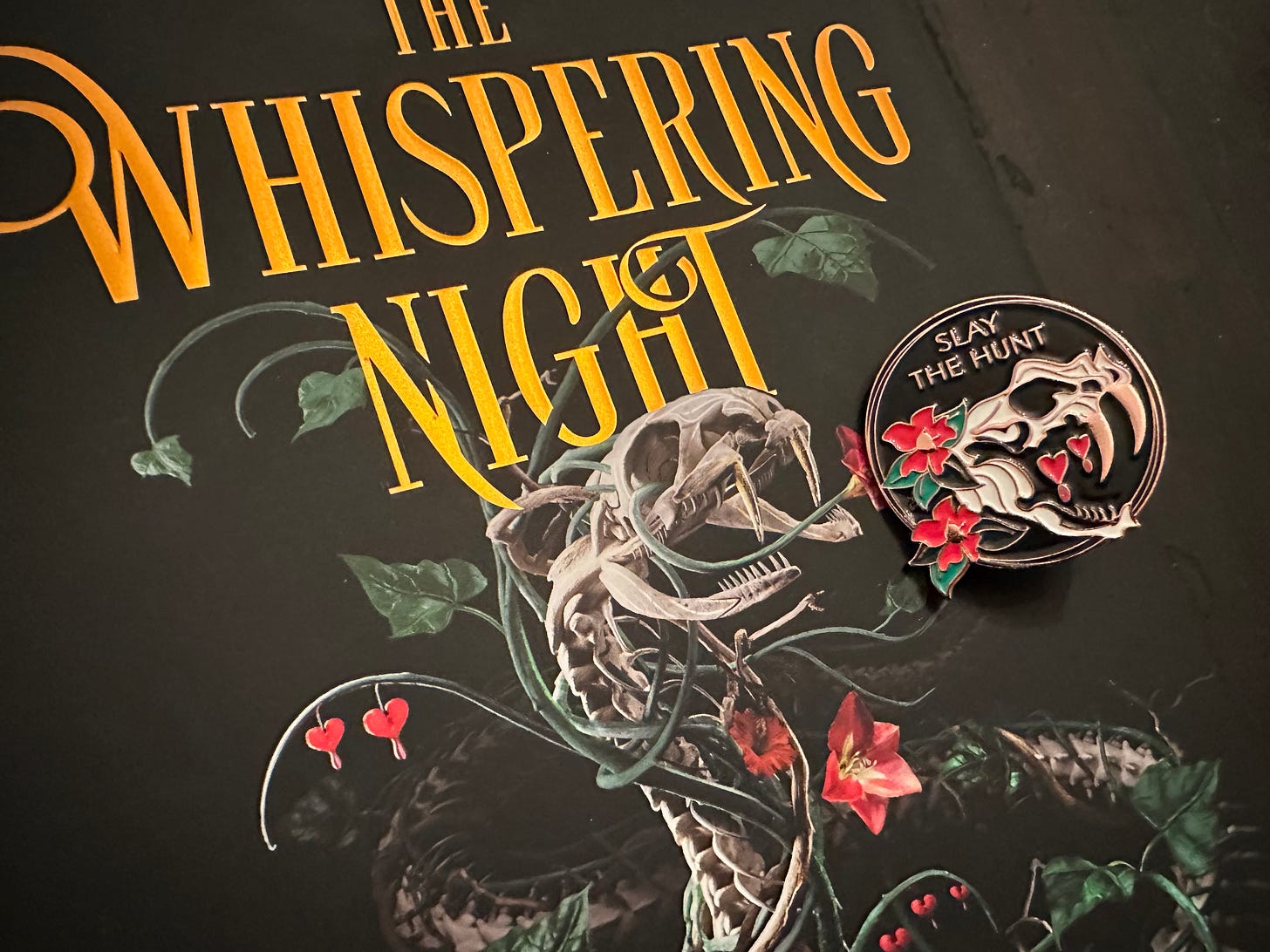 A photo of the enamel pin with a basilisk skull and flowers atop the cover of the Whispering Night