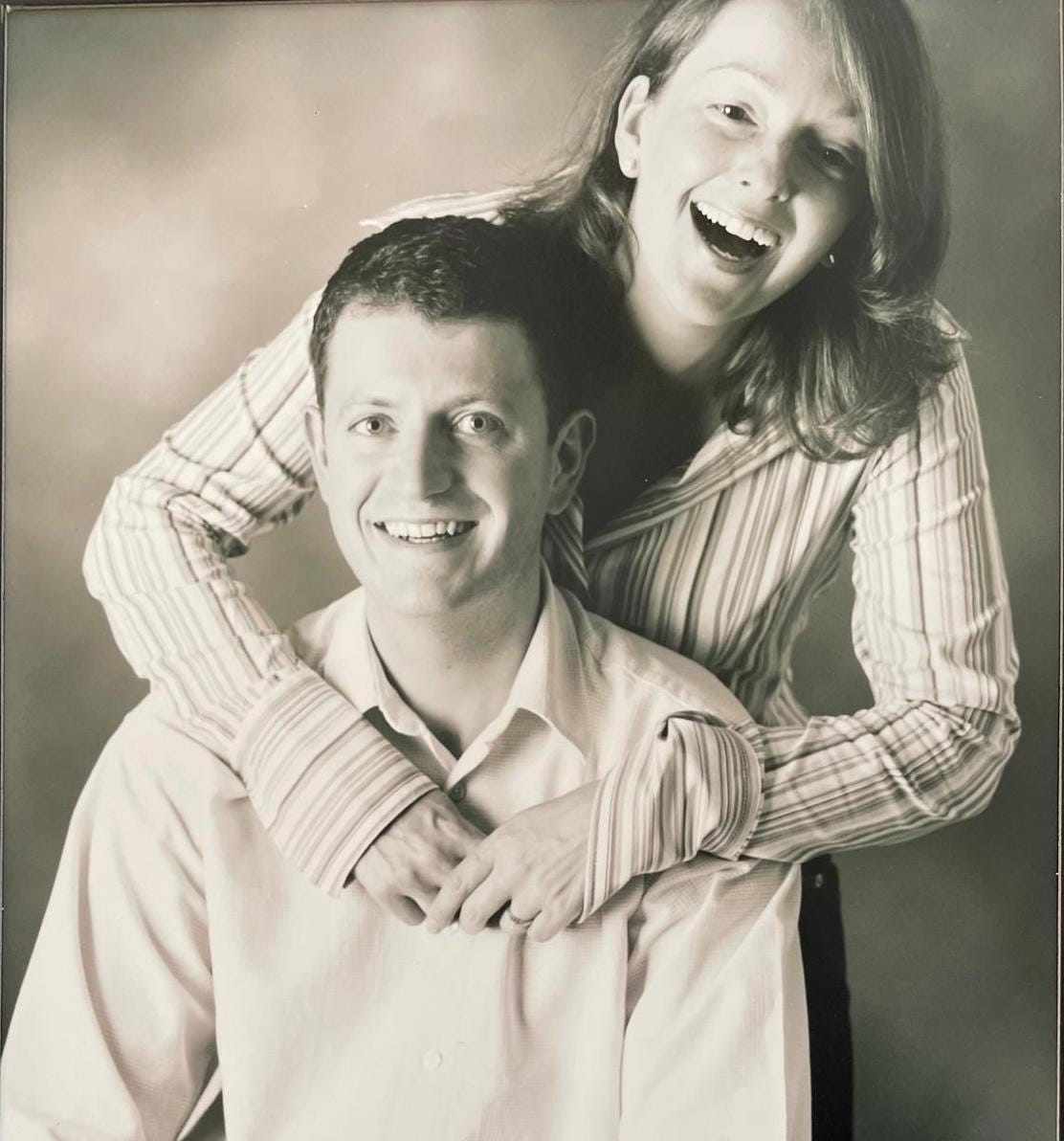 Jane and (husband to be) Jason in a smiley engagement photo pose. Jason is seated with Jane behind with her arms draped around his shoulders Jane and (husband to be) Jason in a smiley engagement photo pose. Jason is seated with Jane behind with her arms draped around his shoulders